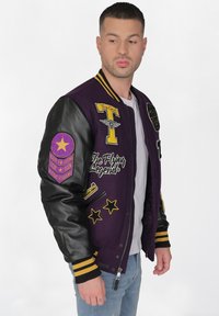 Violetti yliopistojakki mustilla nahkahihansuilla, keltaisilla ja valkoisilla brodeeratuilla tarroilla, raidallisilla hihansuilla ja vetoketjukiinnityksellä.