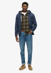 Uomo in felpa blu con zip sopra una camicia verde a quadri, jeans blu arrotolati ai polsini, stivali marroni, cintura marrone e orologio al polso, in piedi di fronte a uno sfondo bianco.