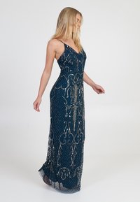 Langes, enges Abendkleid in tiefem Petrol, geschmückt mit silberner Perlenstickerei und aufwendigen Mustern. Es hat dünne Spaghettiträger und einen durchsichtigen Spitzenabschluss.