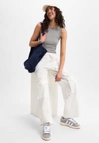 Grijze mouwloze top, witte cargobroek met zakken, grijze sneakers en een marineblauwe schoudertas. Lichte achtergrond met een neutraal podium.