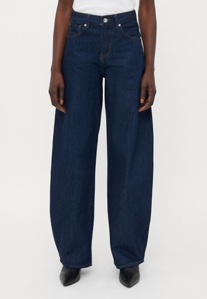 Person trägt dunkelblaue Jeans mit weitem Bein, Fronttaschen und einem knöpfbaren Taillenbund, kombiniert mit schwarzen spitzen Schuhen und einem weißen Oberteil.