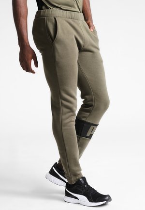 Man draagt olijfgroene slim-fit joggerbroek met zijzakken en zwart-witte sportieve sneakers, staand met handen deels in de zakken.