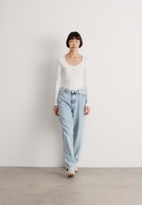 Haut blanc à manches longues avec un décolleté arrondi, associé à un jean bleu clair et ample. Le modèle se tient devant un fond blanc uni.