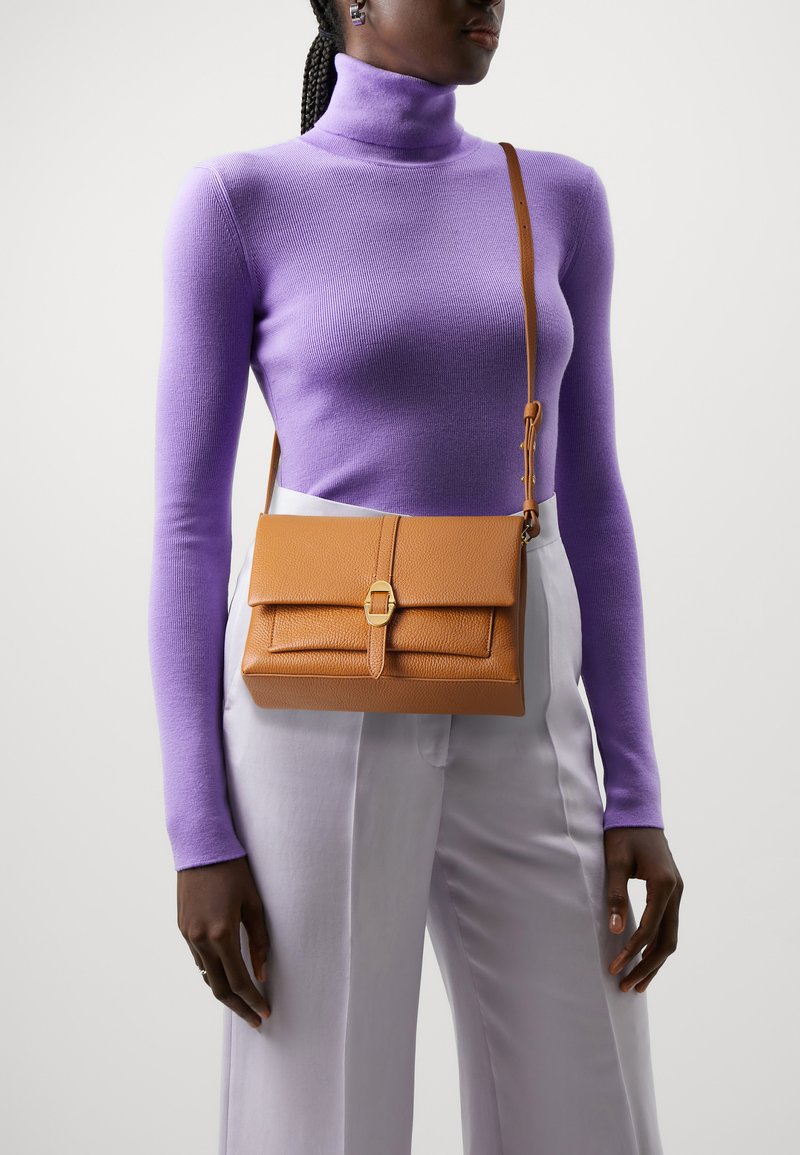 Borsa a tracolla in pelle marrone con design strutturato, patta superiore, accessori in tono oro e finitura testurizzata, abbinata a un dolcevita viola.