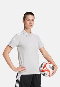 Lichtgrijze poloshirt met een kraag en drie knopen, met witte strepen op de schouders. Zwarte sportshorts. Houdt een meerkleurige voetbal vast.