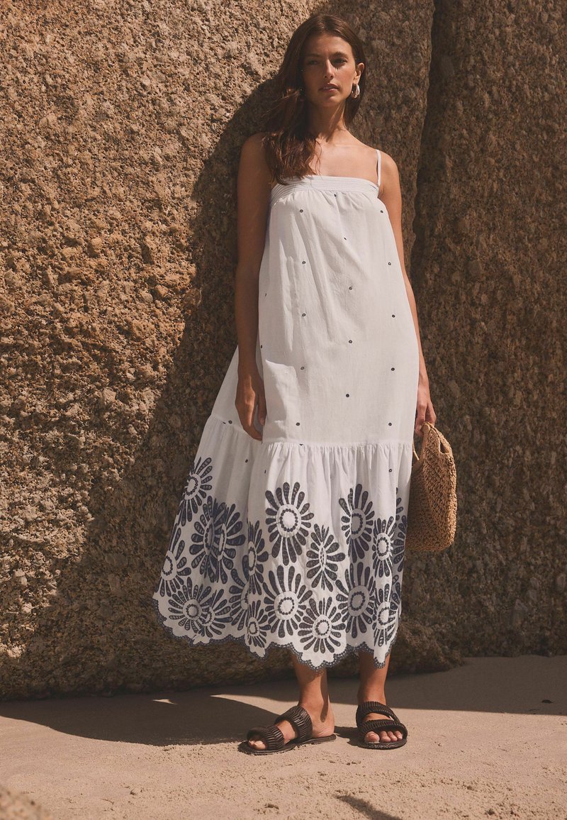 Abito maxi in cotone bianco con ricami floreali blu navy all'orlo, spalline sottili e design a strati. Borsa intrecciata marrone e sandali.
