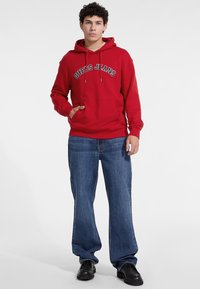 Červená mikina s kapucňou s logom "Guess Jeans" v bielej a námornej farbe, s prednou vreckom a šnúrkami, skombinovaná s modrými voľne strihanými džínsy a čiernymi topánkami.