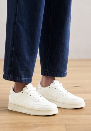 Fehér edzőcipő sima, texturált felülettel, lapos fűzőkkel és platform talppal. Sötétkék, enyhén felgyűrt farmernadrággal viselve.