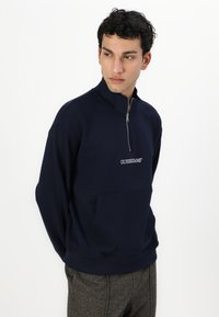 Marineblå fleece-pullover med høj krave, halv lynlås og frontlomme; har broderet logo på brystet og ribbede manchetter.
