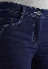 Tmavě modré džíny z denimu s kontrastním béžovým prošíváním, předními kapsami, poutekem na opasek a kovovým knoflíkem se značením na šedém tričku.