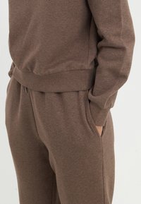 Ensemble de sweatshirt brun et de jogging en tissu doux avec des poignets côtelés et des poches latérales, offrant une coupe décontractée et des coutures minimales.