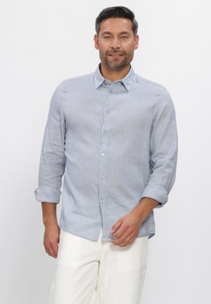 Mann trägt hellblaues Hemd mit aufgekrempelten Ärmeln und weiße Hose, stehend vor einem einfarbigen weißen Hintergrund.