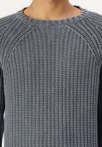 Grå stickad tröja med ribbstruktur, rundad halsringning och raglanärmdesign. Materialet verkar mjukt och medelviktigt.