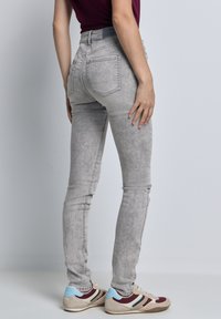 Jeans skinny grises con un acabado desgastado, que presentan bolsillos traseros, una cintura de altura media y un diseño ajustado. Combinados con zapatillas beige y azules.