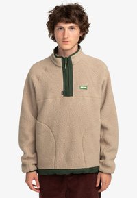 Beige fleece pullover with a green zipper and collar. Features a partial zipper, pockets, and textured fabric with a logo patch.