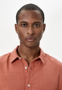 Camisa de botones de color óxido con una tela texturizada, cuello corto y botones blancos. Ajuste estructurado, superficie suave y aspecto natural.