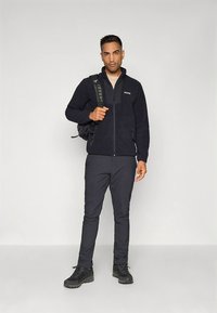 Sötétkék polár dzseki és nadrág, elöl cipzárral és oldalzsebekkel. Fekete hátizsákot visel és masszív fekete cipőt.