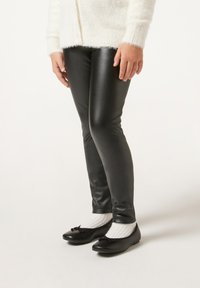 Leggings neri realizzati in materiale lucido con un design aderente, abbinati a ballerine nere con dettaglio a fiocco e calzini bianchi a coste.