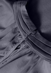 Fialová tkanina s hladkou texturou má plisování a dva shodné knoflíky podél švu, což zdůrazňuje strukturovaný design.