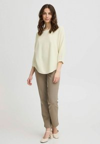 Hellgelbe, strukturierte Bluse mit Dreiviertelärmeln, kombiniert mit beigen, taillierten Hosen und hellen, hochhackigen Schuhen, vor einem neutralen Hintergrund stehend.