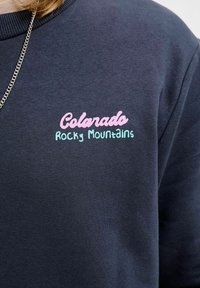 Sudadera azul oscura con "Montañas Rocosas de Colorado" bordado en rosa y verde azulado. Presenta un cuello de canalé y una textura suave.