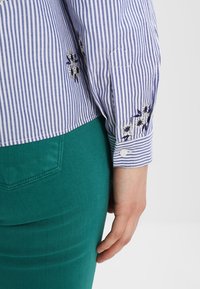 Chemise à manches longues rayée bleu et blanc avec des fleurs brodées près du poignet et dans le dos, portée avec un pantalon vert sarcelle.