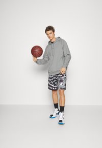 Felpa grigia, pantaloni corti neri e grigi a fantasia con logo dei Raiders. Scarpe sportive bianche e nere, con in mano un pallone da basket, in piedi su una superficie semplice.
