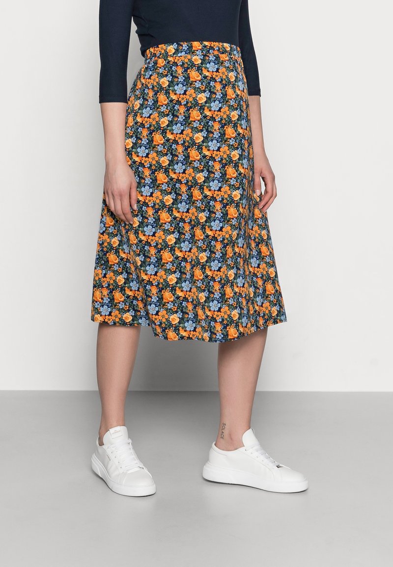 Žena v sukně po kolena s květinovým vzorem v oranžové a modré barvě, v bílých teniskách a černém topu s dlouhým rukávem, stojící na šedé podlaze.