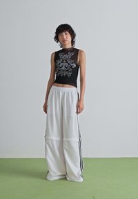 Young person stands on green floor against plain wall, wearing black sleeveless top with silver design and oversized white track pants with stripes.