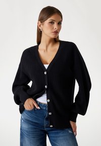 Black knitted cardigan with a V-neck, oversized sleeves, and decorative button details. Paired with blue distressed jeans.
