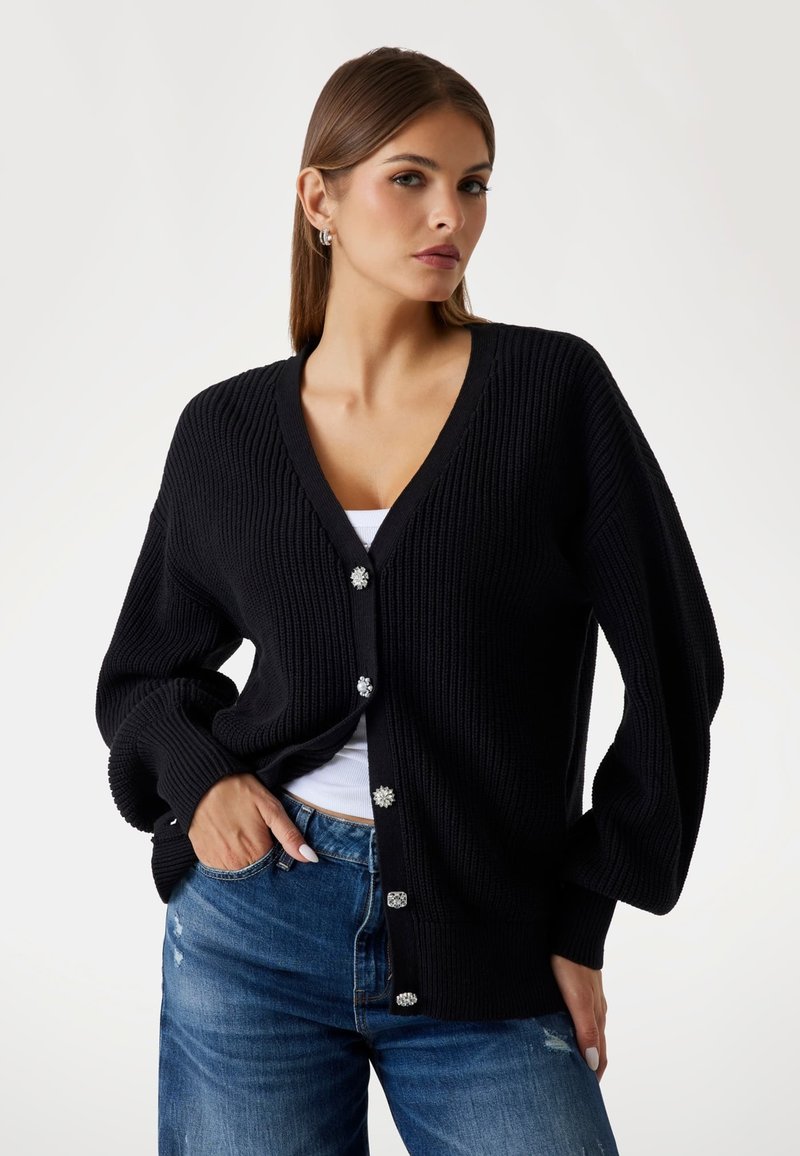 Black knitted cardigan with a V-neck, oversized sleeves, and decorative button details. Paired with blue distressed jeans.