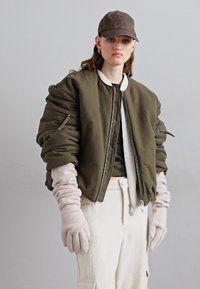 Olīvu zaļš bombera jakas ar quilted detaļām, balta rāvējslēdzēja, krēmkrāsas cimdiem un saskaņotām biksēm; strukturēts, ikdienišķs dizains.