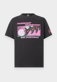 Μαύρο μπλουζάκι Nike με ροζ γραφικό από ποδοσφαιρικά παπούτσια που κλωτσούν μια μπάλα ποδοσφαίρου, κείμενο «Φιλικό Τουρνουά» και «Nike Sportswear» στο μπροστινό μέρος.