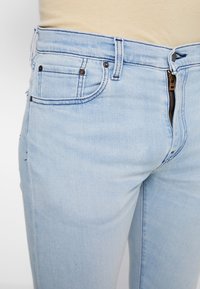 Světle modré džíny z denimu mají zipový poklopec a pět kapes s kontrastním prošíváním a kovovými nýtky v blízkosti kapes.