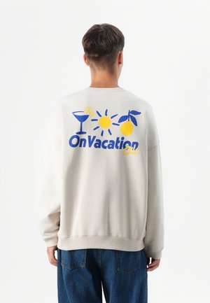 A person in a light gray sweatshirt with a colorful vacation-themed design on the back, facing away against a white background.