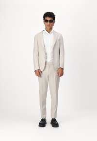 Costum bej deschis cu sacou structurat și pantaloni drepti. Cămașă albă dedesubt. Model purtând ochelari de soare negri și pantofi închiși la culoare.