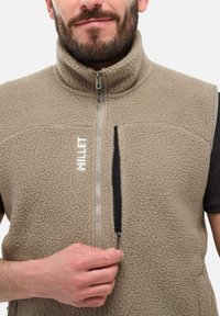 Mann trägt eine beigefarbene Fleeceweste mit einem Reißverschlusstaschen, hält den Reißverschluss der Tasche fest, ein Teil seines Gesichts und seines Bartes ist sichtbar.