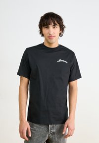 Camiseta de algodón negra con cuello redondo, mangas cortas y un logo blanco de "ellesse" en el pecho, combinada con unos jeans claros desgastados.