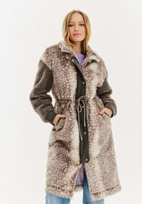 Žena v dlouhém kabátu z umělé kožešiny s páskem, se skvrnitým vzorem a texturovanými rukávy, stojící s rukama v kapsách na jednobarevném pozadí.