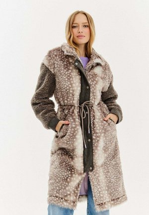 Kobieta w długim, futrzanym płaszczu z paskiem, w plamisty wzór i teksturowanymi rękawami, stoi z rękami w kieszeniach na jednolitym tle.