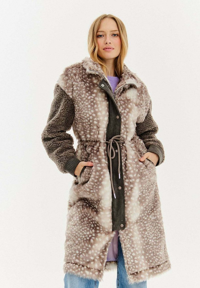 Žena v dlouhém kabátu z umělé kožešiny s páskem, se skvrnitým vzorem a texturovanými rukávy, stojící s rukama v kapsách na jednobarevném pozadí.