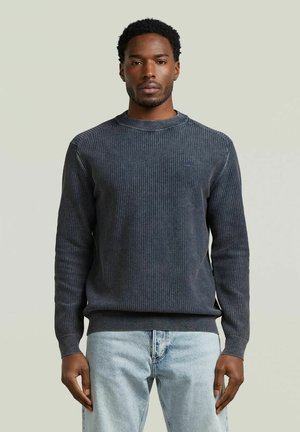 Uomo che indossa un maglione a coste grigio scuro e jeans azzurri, in piedi davanti a uno sfondo chiaro e uniforme, rivolto in avanti.