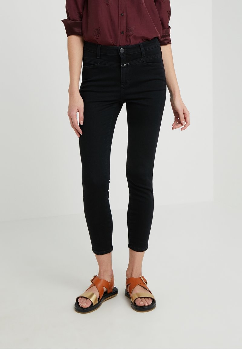 Personne portant un jean skinny noir, une chemise bordeaux à manches longues et des sandales ouvertes marron et dorées, debout sur un sol blanc.