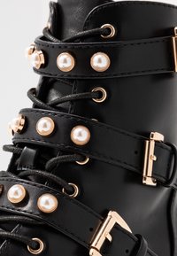 Černé kožené boty s zlatými akcenty, perlovými nýty a křížovými pásky. Texturovaný povrch a kontrastní černé tkaničky.