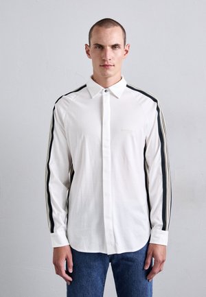 Hombre vestido con una camisa de manga larga blanca y cuello, con rayas negras y beige en las mangas, y jeans azules, de pie contra una pared clara.