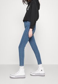 Jeans en denim bleu skinny avec une taille haute, associés à un sweatshirt à capuche noir et des baskets montantes blanches avec des rayures noires.
