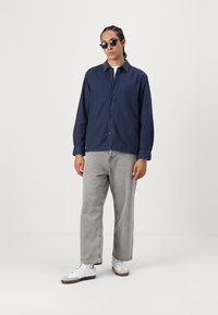 Navy blue button-up shirt, light grey loose jeans, white sneakers with grey accents, and dark sunglasses against a plain background.