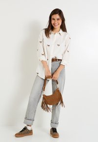 Chemise blanche avec des motifs de chats bruns, jean gris taille haute, sac à main à franges marron et baskets à pois. Tenue décontractée avec différentes textures.