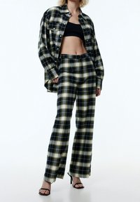 Schwarz-gelb karierte Bluse und Hose aus weichem Stoff, mit lockerer Passform und weitem Bein. Schwarze High Heels vervollständigen das Outfit.