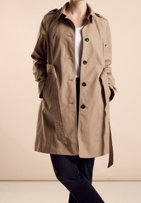 Béžový trenčkot s páskem, zapínáním na knoflíky, ramenními klapkami a bočními kapsami, vyrobený z hladké látky, nošený přes bílé tričko a tmavé džíny.